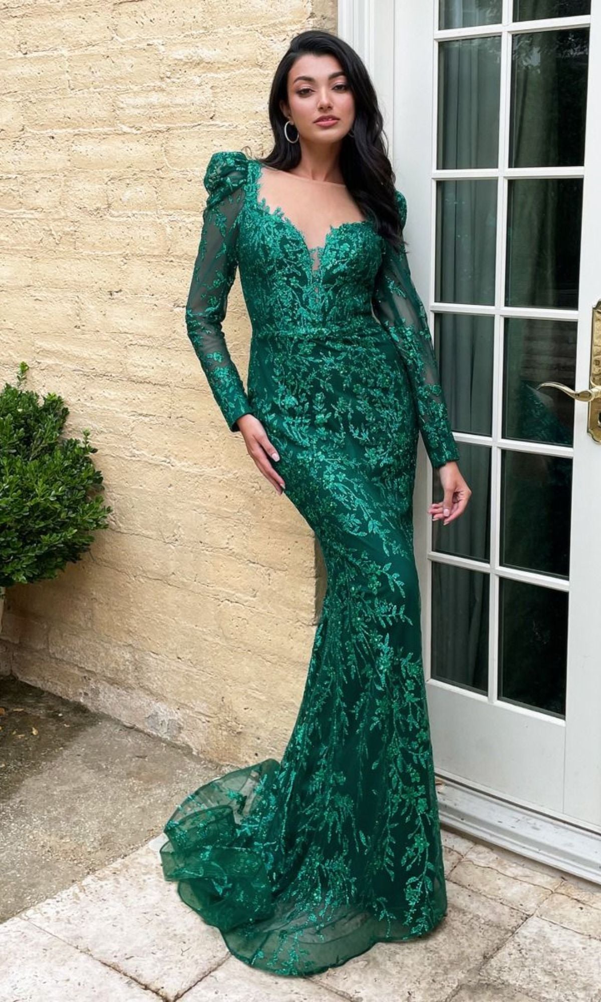Woman modeling the Long Sleeve Long Glitter-Lace Prom Dress CD989, a fitted, green mermaid gown with floral lace details and glitter, standing by a cream brick wall and glass door.